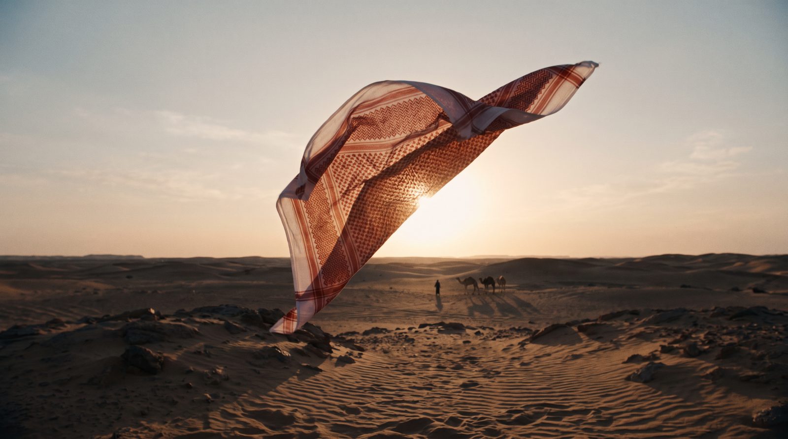 الشماغ يتحرك مع الريح قدام قرص الشمس، وخلفه رجل وقافلة جمال في الأفق.