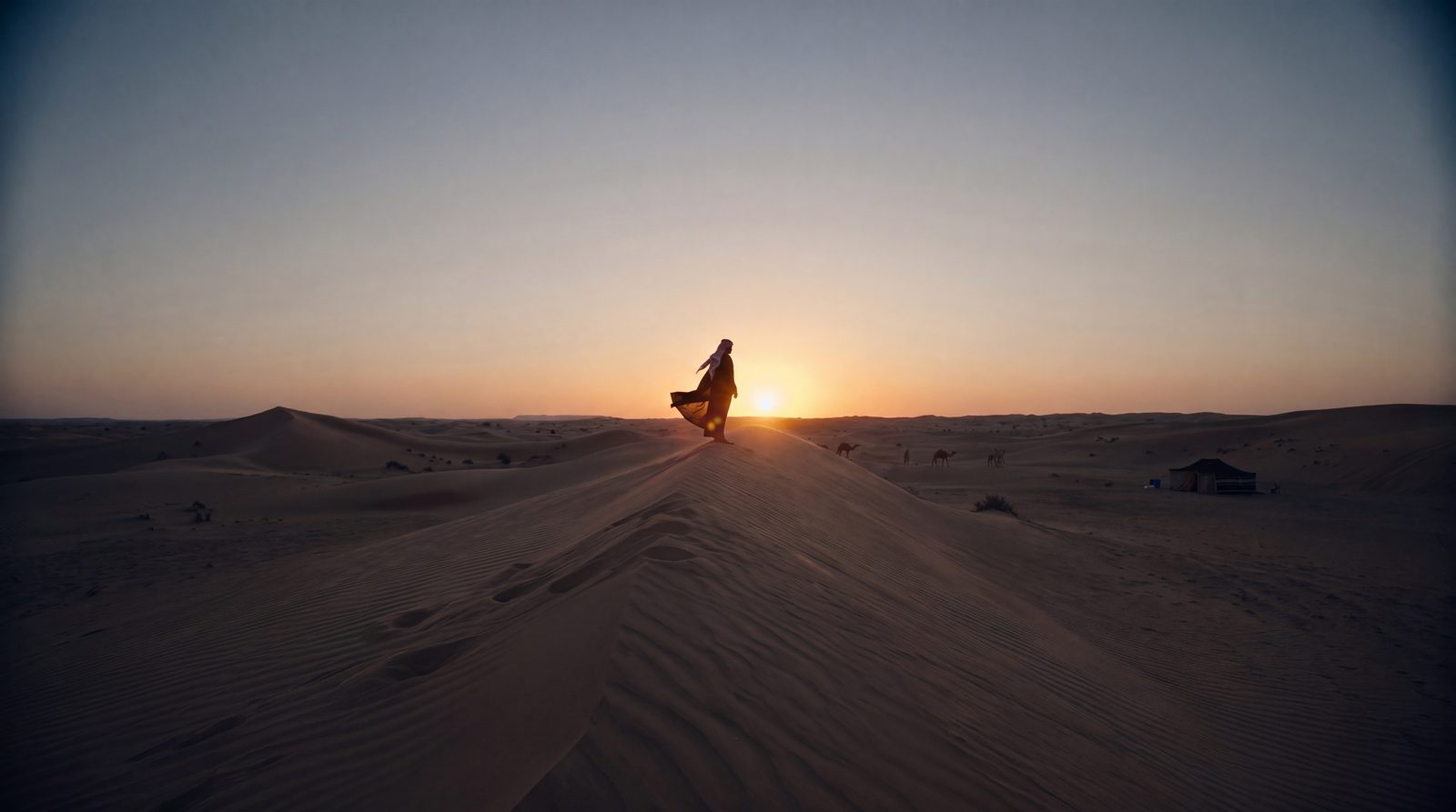 رجل على قمة الكثيب مقابل الغروب، والريح تشد عباته.