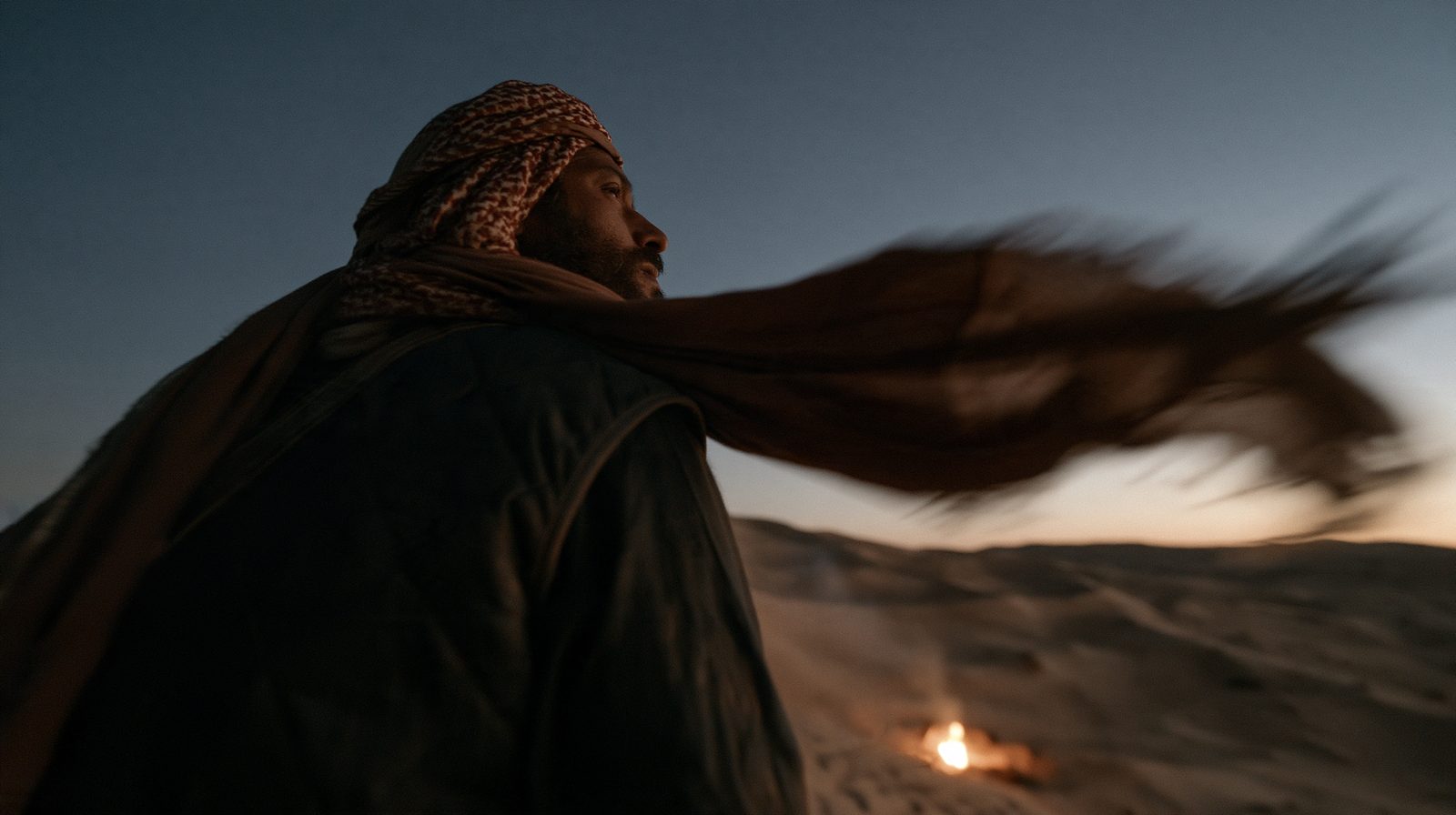 زاوية واطية: رجل يتلحف بشماغه والريح تشده، ونار على الكثيب وراه.